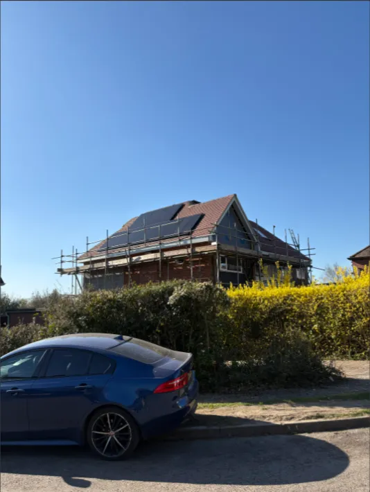 House with solar panels on its roof, scaffold around it, and a car parked nearby in sunny weather